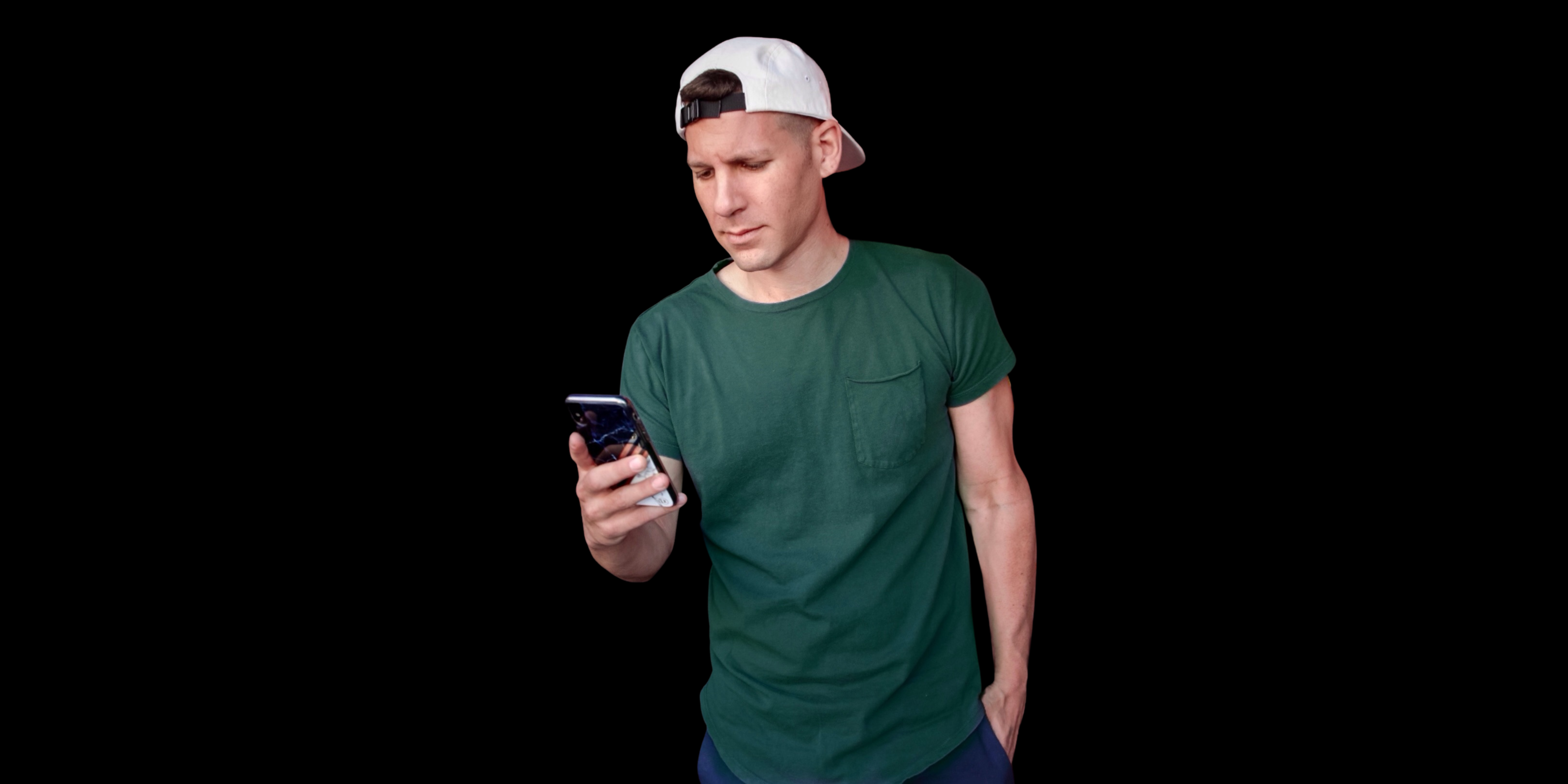 Man wearing a green t-shirt and white backwards cap looks at his phone with a neutral expression, standing against a black background.