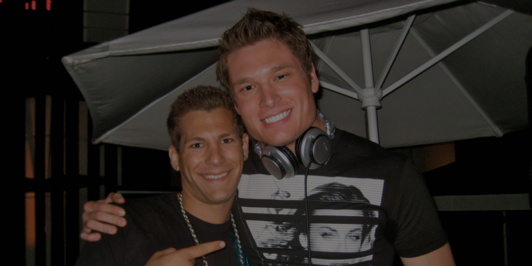 Two young men smile at the camera; one wears a necklace and points, while the other has headphones around his neck and a graphic t-shirt. An outdoor umbrella is in the background.