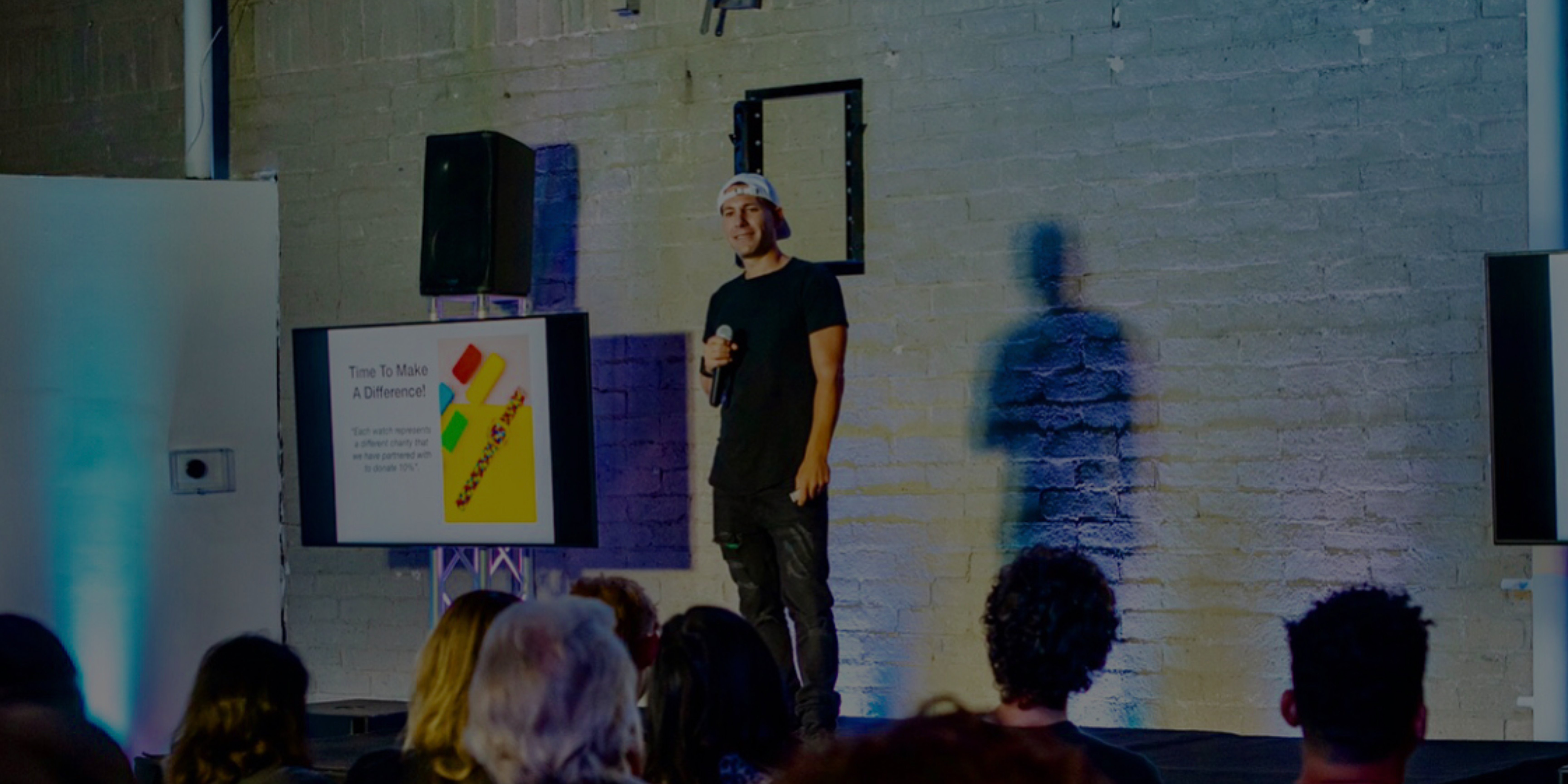 A man in casual clothing stands on stage holding a microphone, presenting to an audience; a screen beside him displays a slide titled "Time to Make A Difference!.