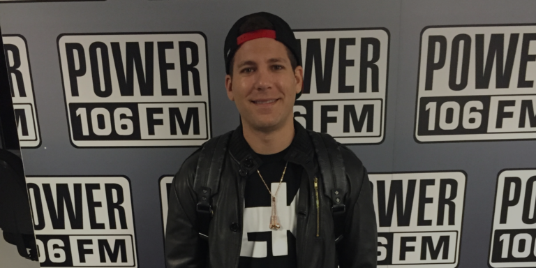 A person in a black jacket and backwards cap stands in front of a wall with multiple Power 106 FM logos.