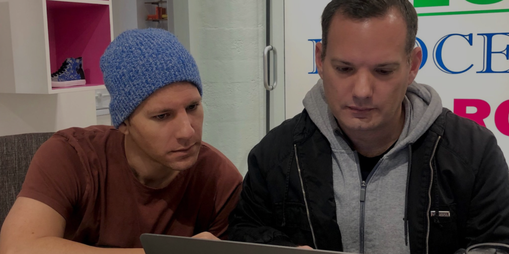My Brother Brad. Two men sit closely together, focused on a laptop screen. One wears a blue knit beanie and a brown shirt, the other wears a gray hoodie under a black jacket.