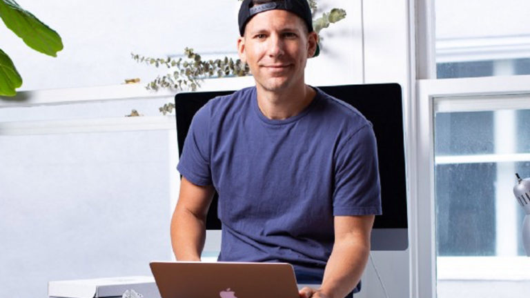 A man in a blue t-shirt and backwards cap sits at a desk with a laptop, in front of a large monitor and window, looking at the camera.