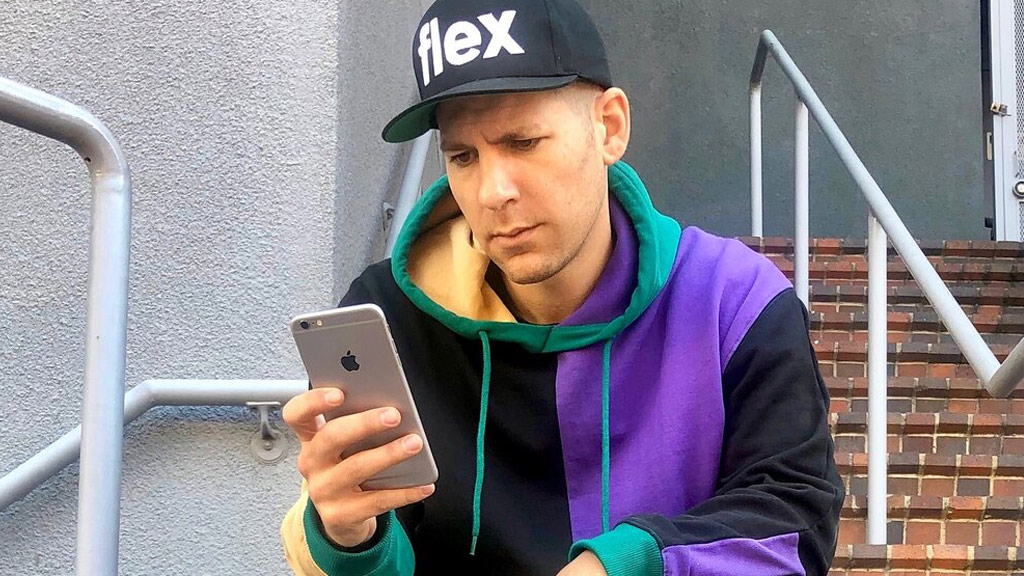 9 Simple Ways To Improve Your Chances of Success...Sitting Down A man wearing a "flex" cap and a colorful hoodie sits on outdoor steps, looking at a smartphone.