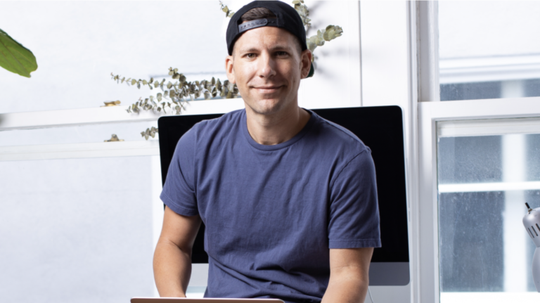 A man in a backwards black cap and blue t-shirt sits indoors holding a laptop, with a computer monitor and large windows in the background.