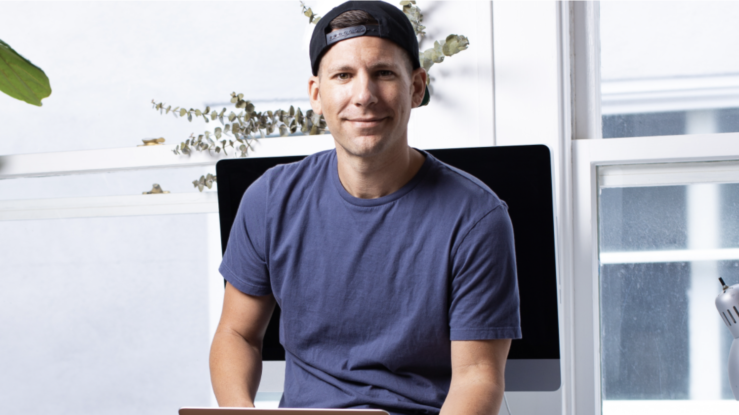 A man in a backwards black cap and blue t-shirt sits indoors holding a laptop, with a computer monitor and large windows in the background.