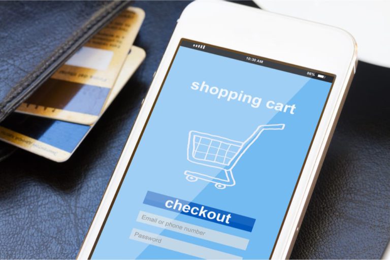 A smartphone displaying a shopping cart and checkout screen is placed next to a wallet containing several credit cards.