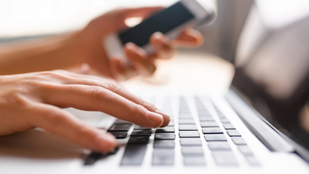 Person typing on a laptop keyboard with one hand while holding a smartphone in the other hand.