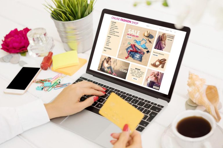 A person shops for clothes on an online fashion website using a laptop, holding a credit card, with a phone, coffee, and stationery on the desk.