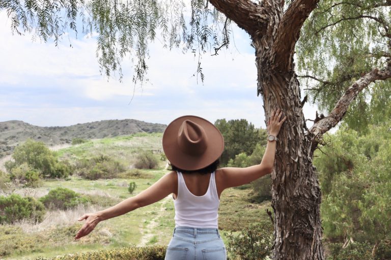 A person wearing a brown hat, white tank top, and jeans stands with arms outstretched, facing a tree and a green landscape under a partly cloudy sky.