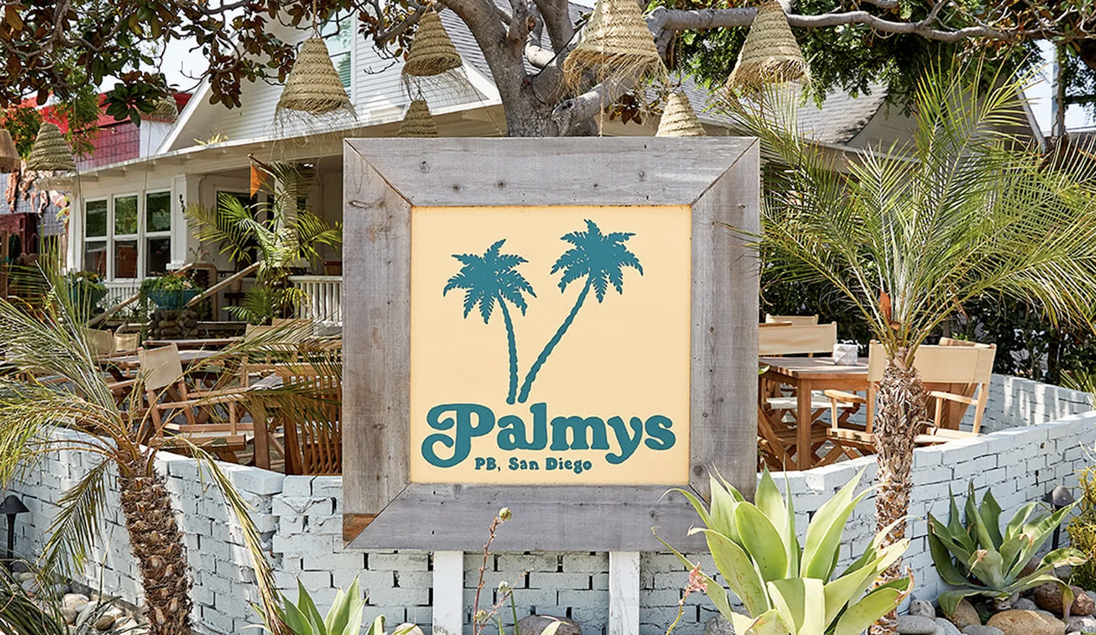A wooden sign with two teal palm trees and the text "Palmys PB, San Diego" stands in front of an outdoor seating area with tropical plants and wooden tables.