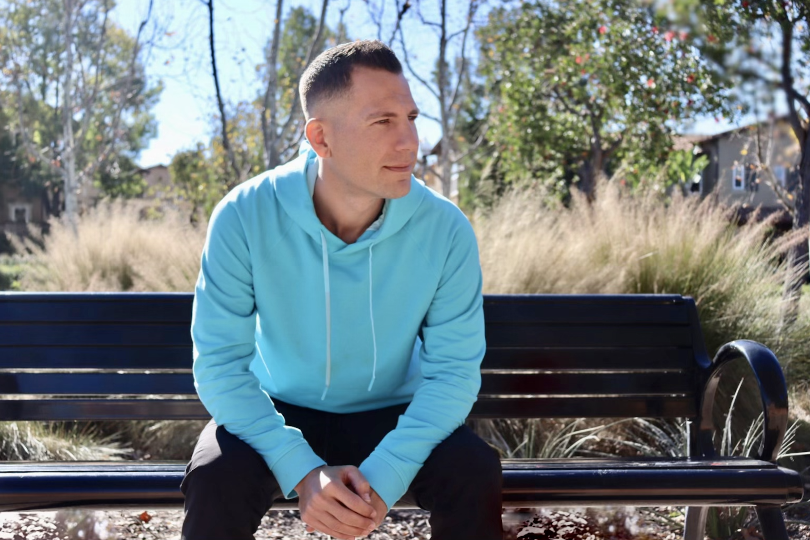 A man in a light blue hoodie sits on an outdoor bench, looking to his left, with trees and bushes in the background.