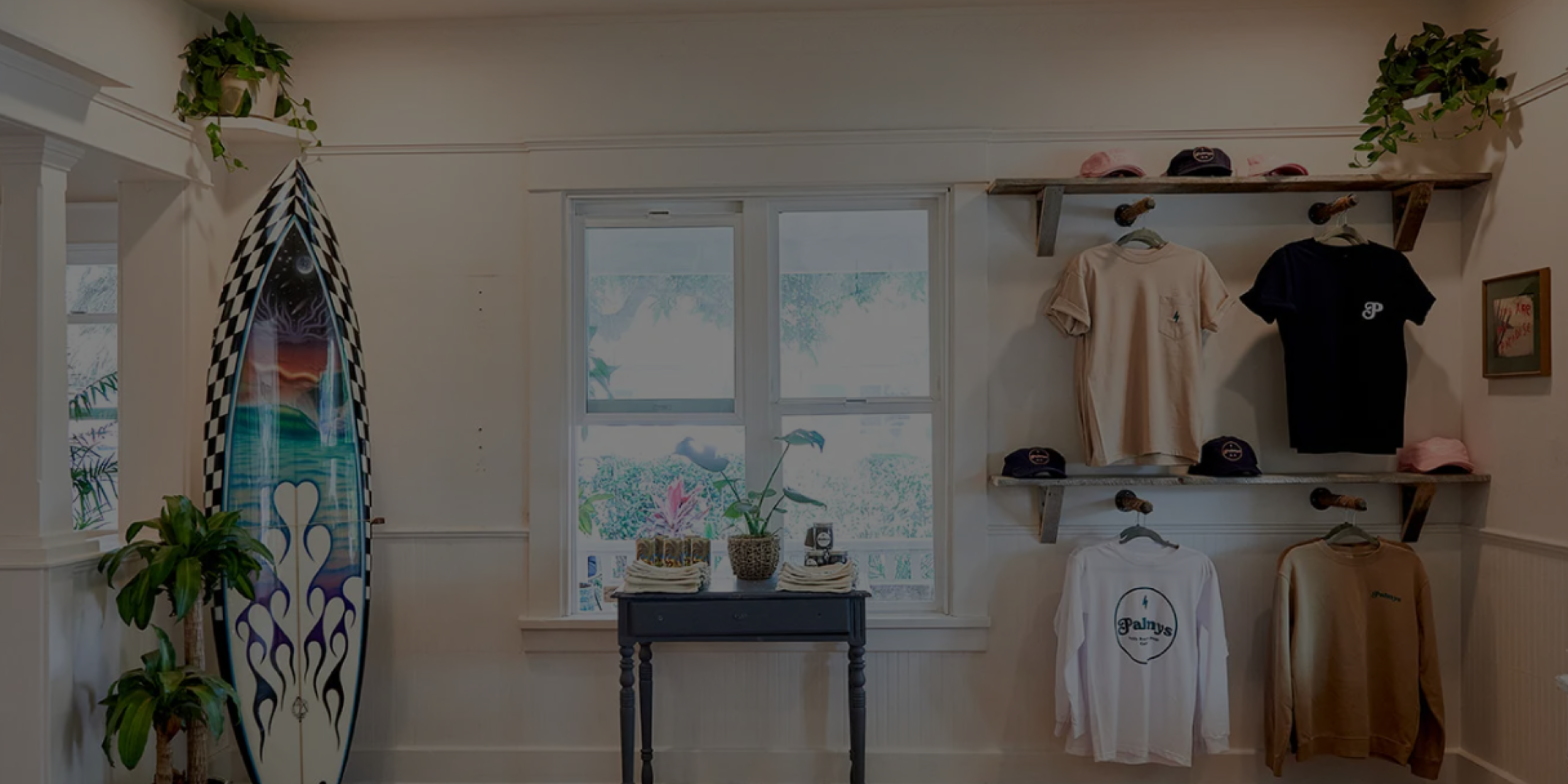 A small display area with a surfboard, potted plants, a table, and wall shelves holding hats and shirts in a room with white walls and a window.