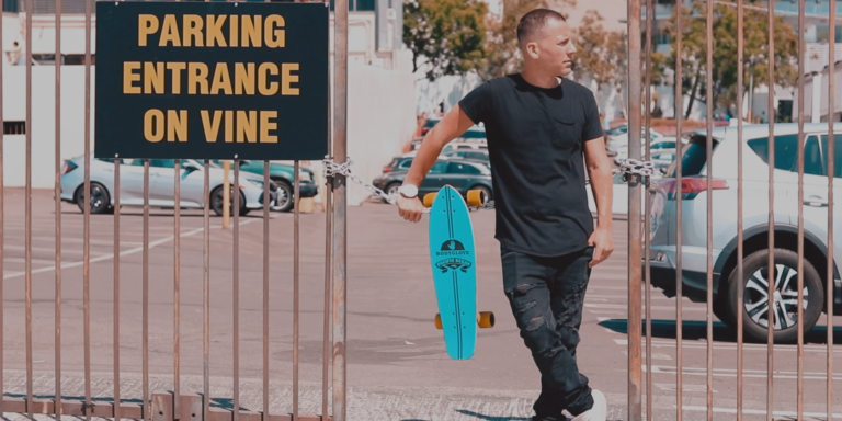A person holding a blue skateboard walks through a gate next to a sign reading "Parking Entrance On Vine" in a parking lot.
