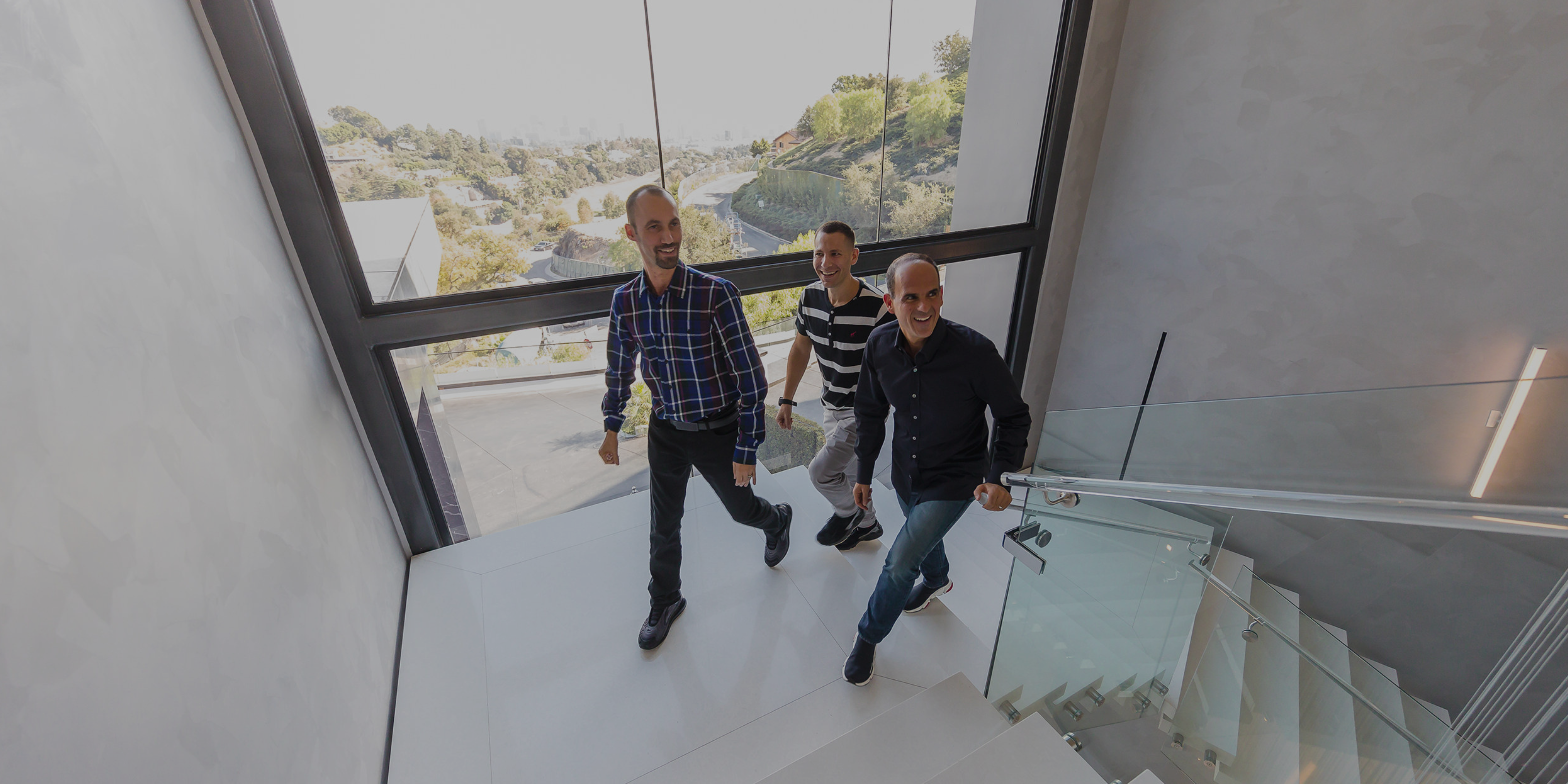 Three men walk up a modern indoor staircase with large windows overlooking a landscape with trees and buildings.