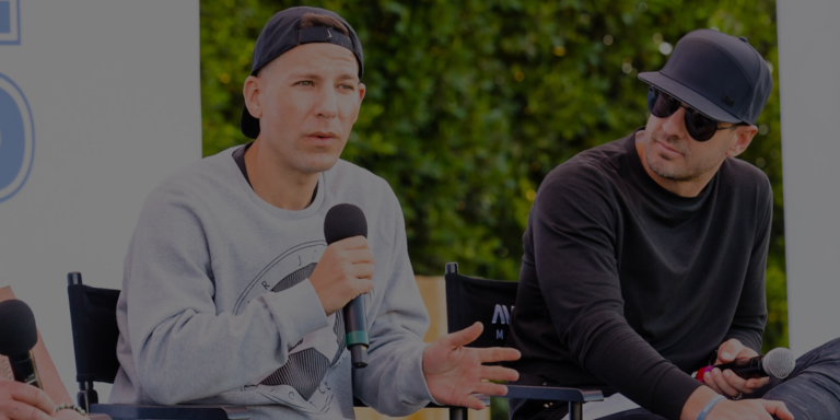 Two men sit outdoors, one speaking into a microphone and gesturing with his hand, while the other listens. Both wear casual clothing and hats. Green foliage is visible in the background.