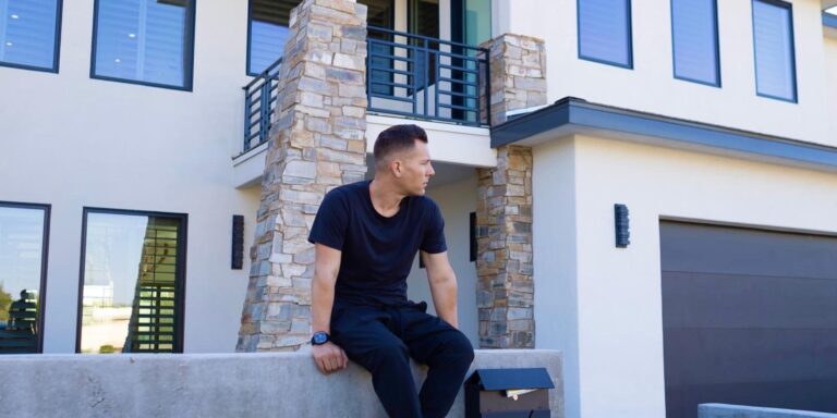 A man in black clothing sits on a concrete wall in front of a modern house with large windows and stone accents, looking to his left.