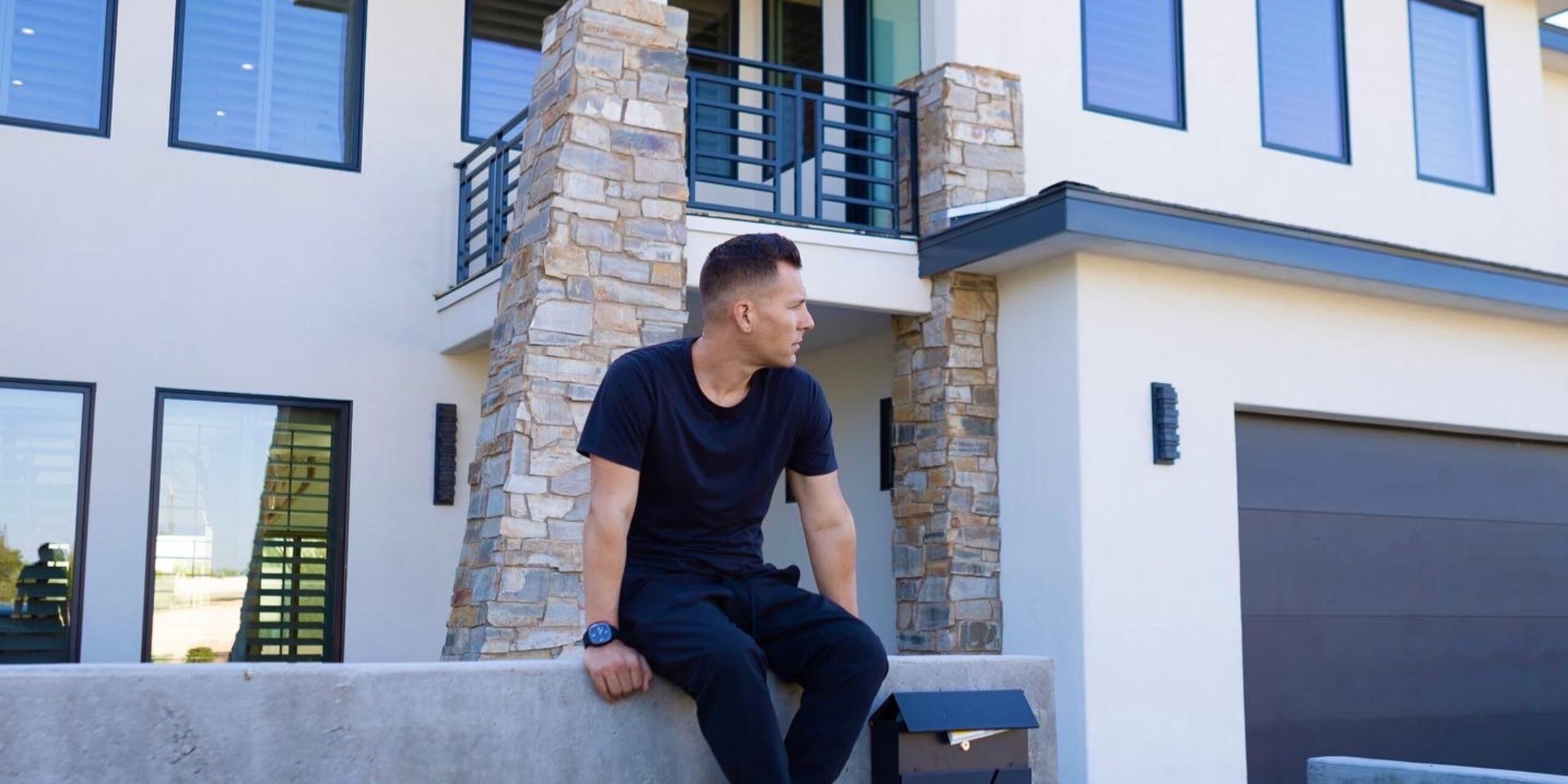 A man in black clothing sits on a concrete wall in front of a modern house with large windows and stone accents, looking to his left.