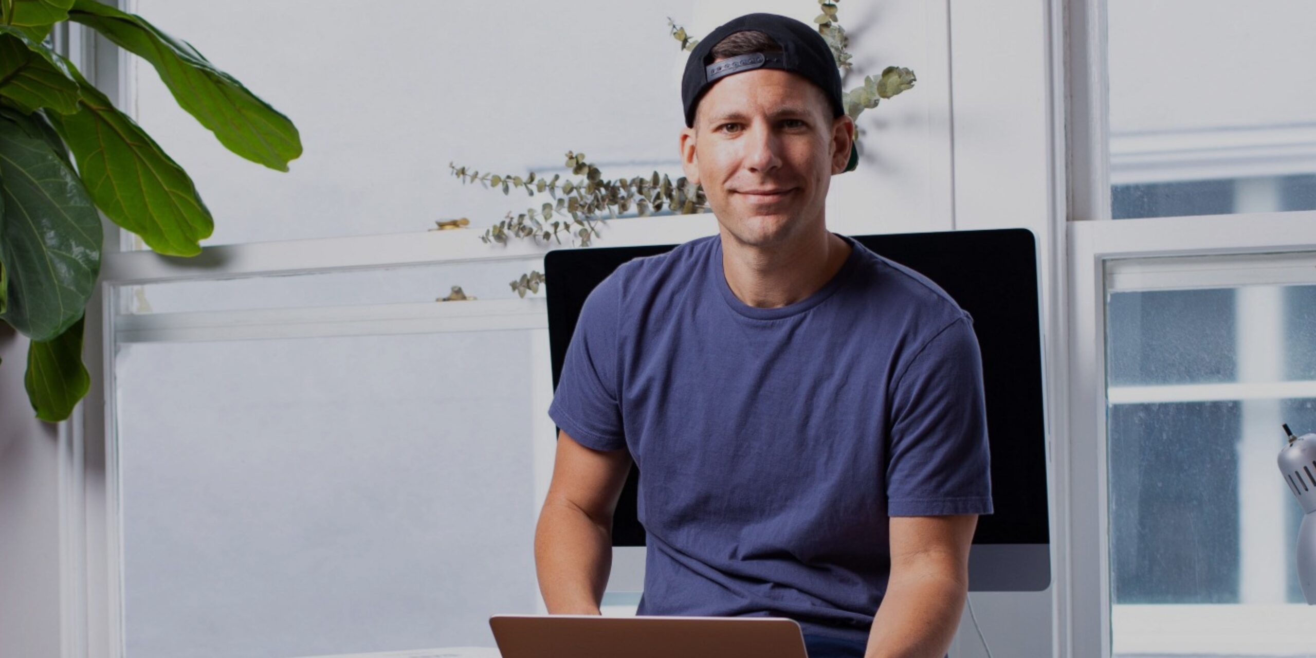Man in a blue t-shirt and backwards cap sits at a desk with a laptop, in front of a large monitor, with plants and a window in the background.