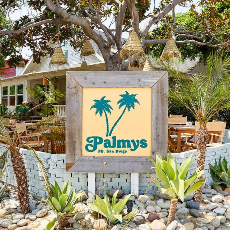 A wooden-framed sign with two palm trees and the text "Palmys P.B. San Diego" stands in front of an outdoor patio with tables, plants, and hanging wicker lights.