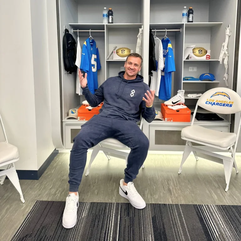 A man in a navy tracksuit sits smiling and gesturing in a locker room with Los Angeles Chargers gear and uniforms displayed behind him.