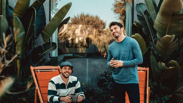 two men are outdoors in front of a large window surrounded by tropical plants; one is sitting on an orange chair holding a mug, and the other is standing beside him, also holding a mug.